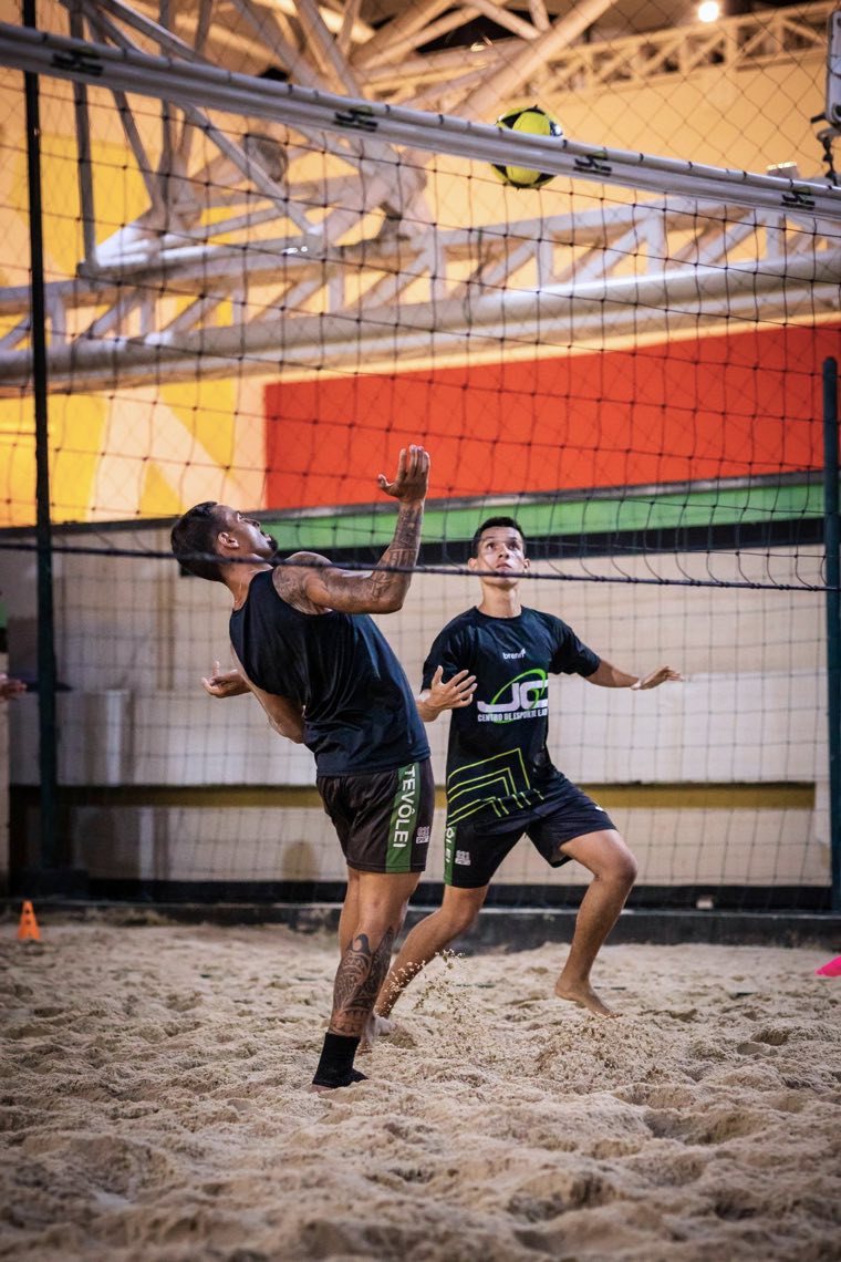 Jogador de futevôlei atacando a bola no alto junto à rede, em quadra de areia.