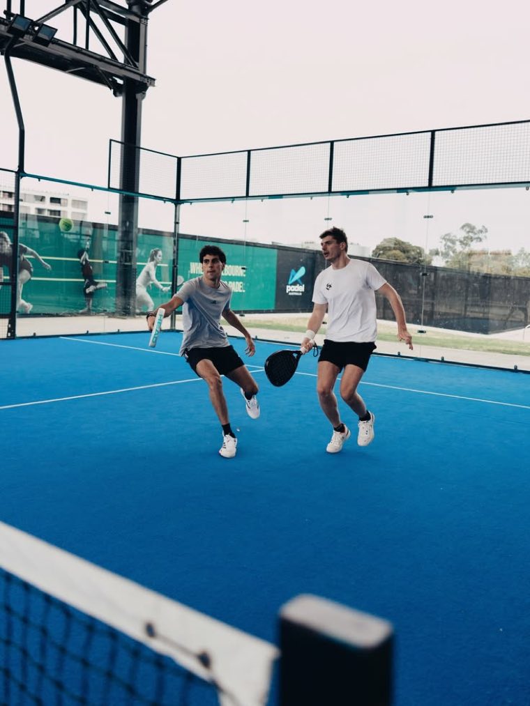 Dois jogadores de padel disputando um ponto em quadra azul ao ar livre.