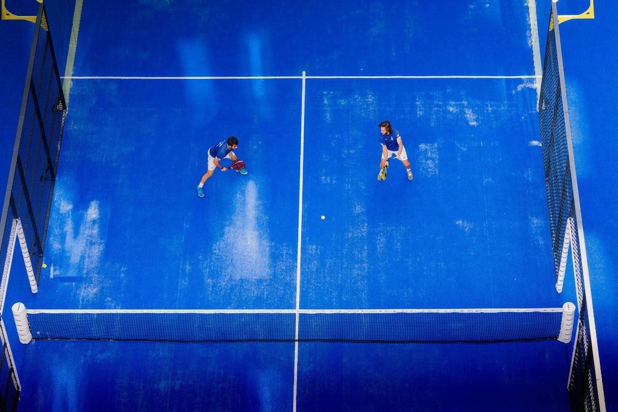 Vista aérea de uma quadra de padel coberta durante um ponto, com dois jogadores posicionados.